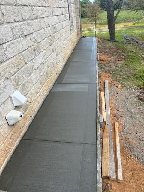 Freshly finished concrete sidewalk with scored lines bordering a light tan stone masonry building wall.