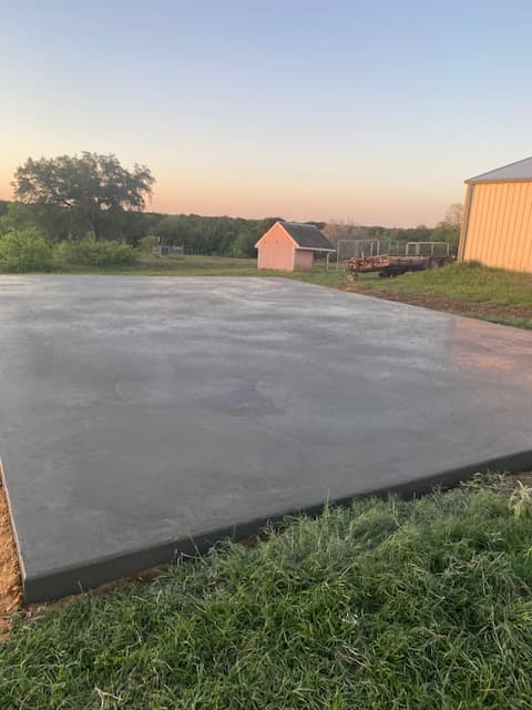 Freshly poured concrete pad in a grassy field next to a yellow barn at sunset.