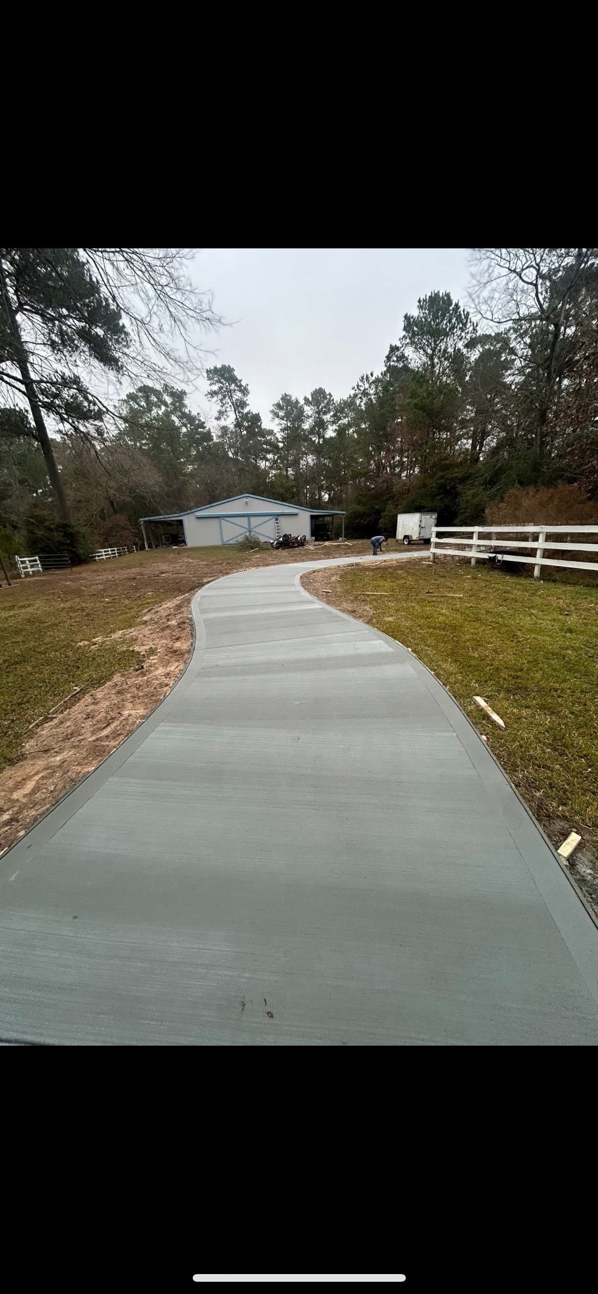 A long, winding concrete driveway with a broom finish leading toward a barn and trees.