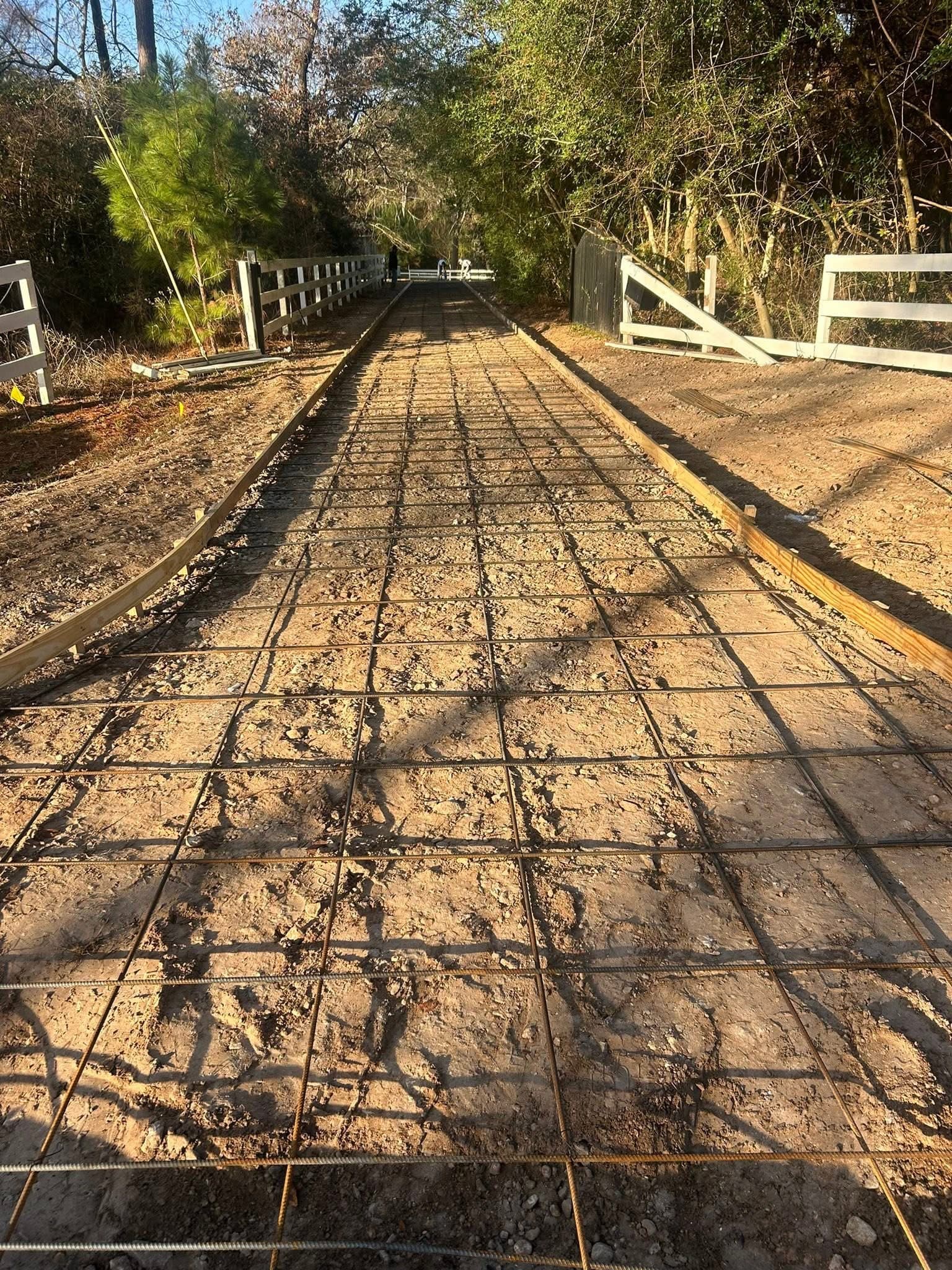 Dirt path with wooden forms and rebar grid prepared for concrete pouring between white fences.