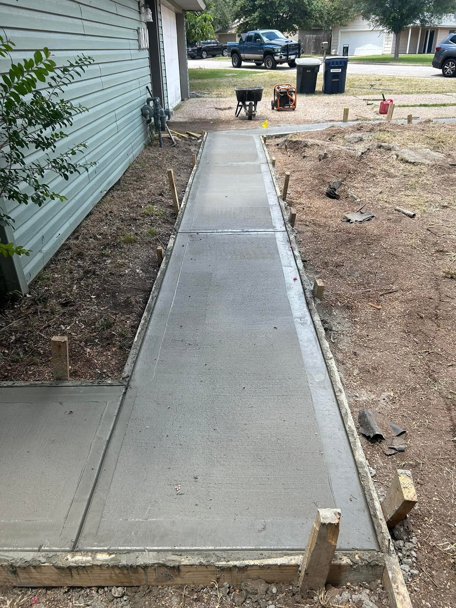 Freshly poured concrete walkway with wooden forms running alongside a house with light green siding.